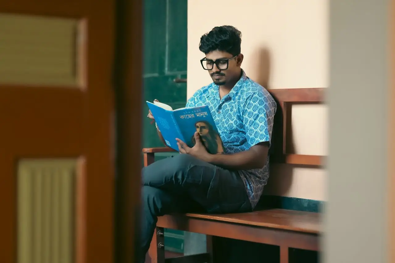 A young man sitting on a bench reading a book indoors. Calm and focused study environment.